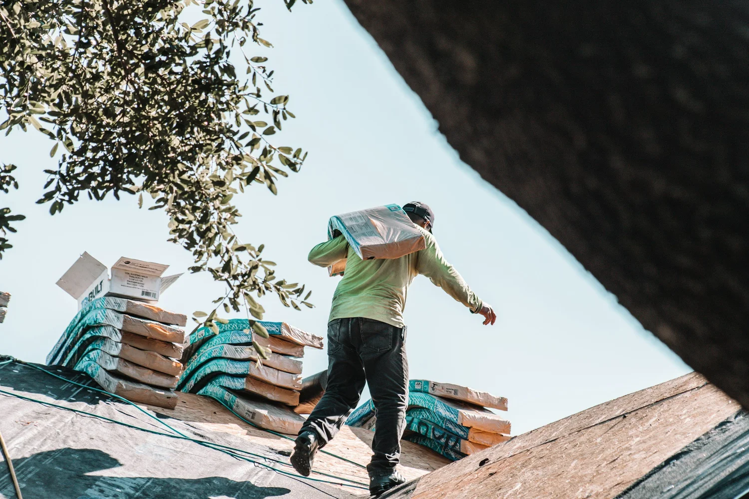 Worker carrying roofing materials on a steep roof with trees in the background.