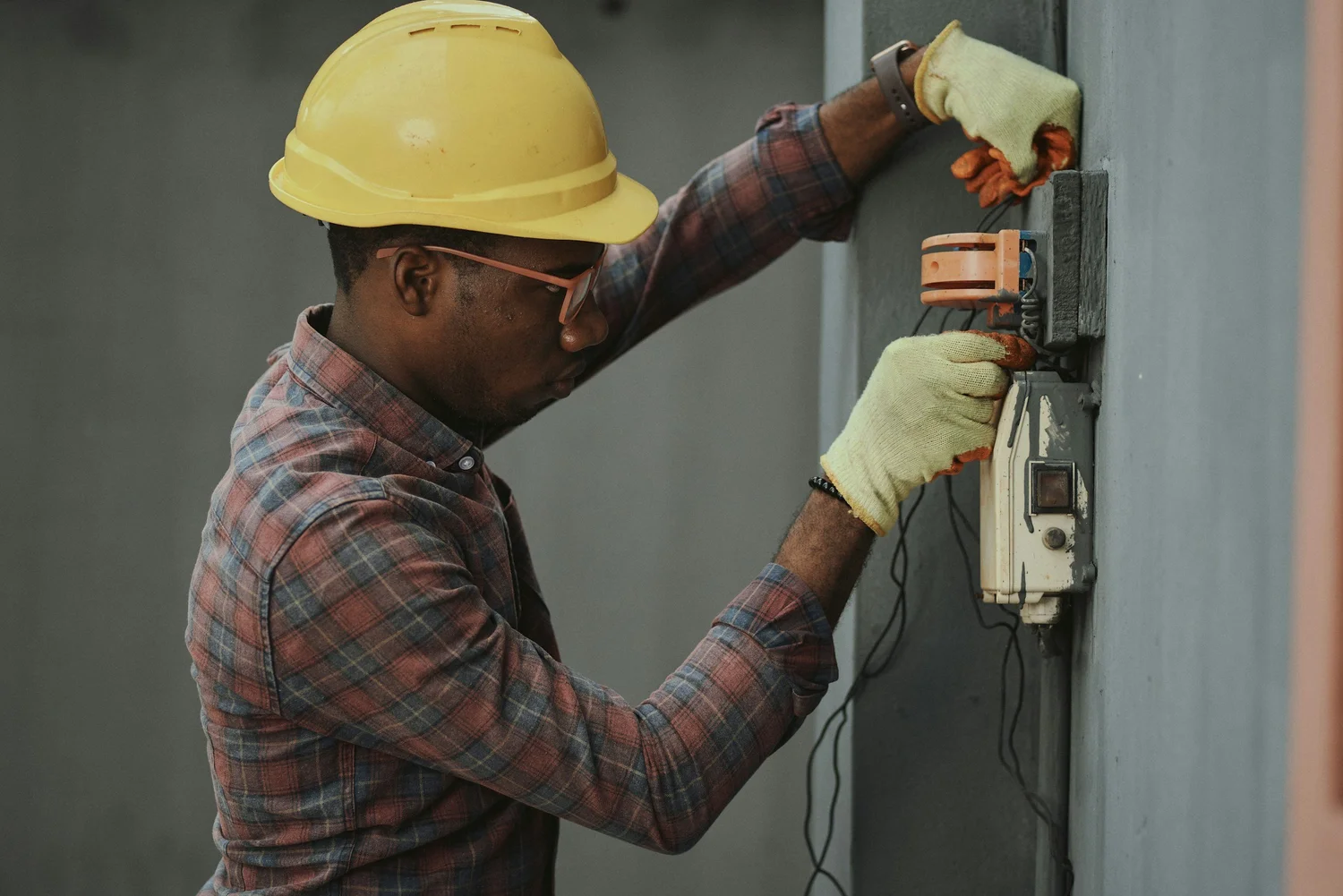 Electrician wearing a yellow hard hat and gloves working on an electrical panel mounted on a wall.